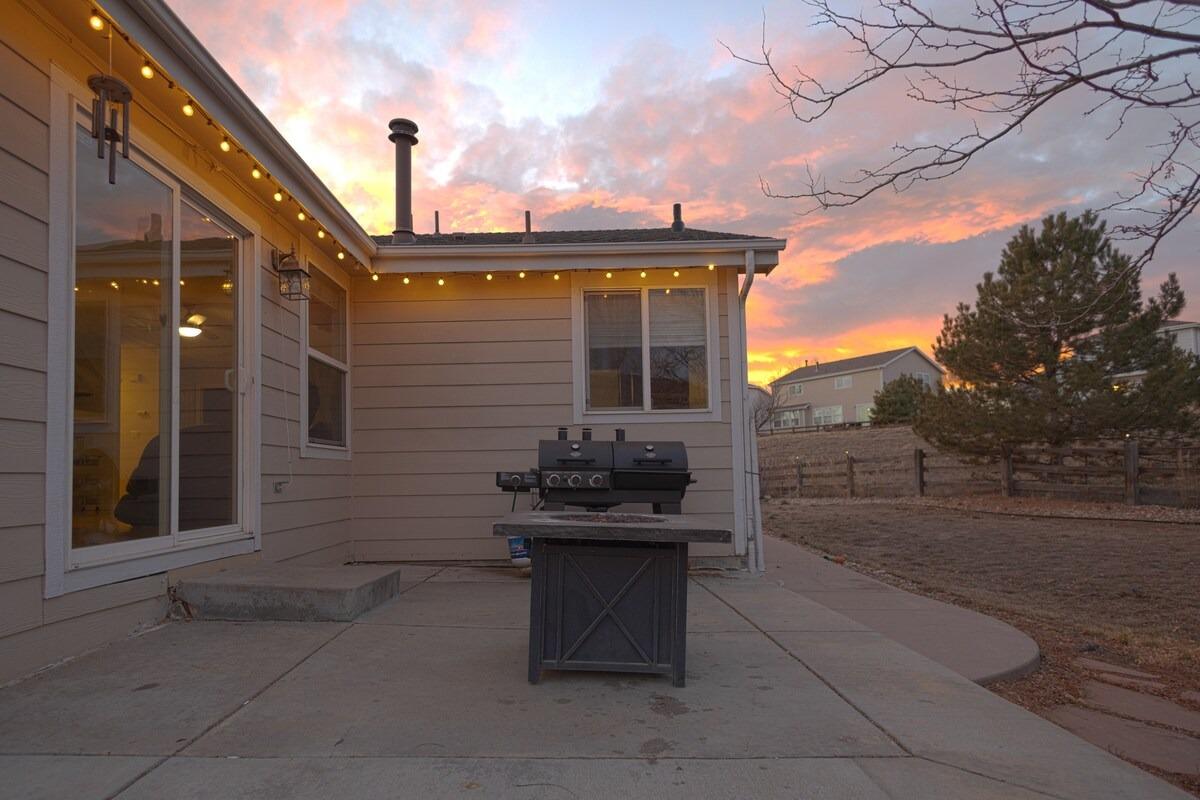 Back patio with string lights and a grill at dusk, fenced yard and pine trees beyond